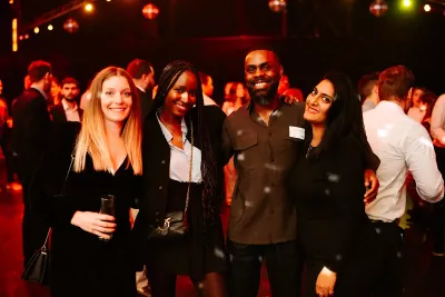 Four people smiling and posing together at a party with warm lighting and a crowd in the background.