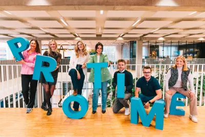 Group of people in an office holding large blue letters spelling 'PROTIME' and smiling at the camera.