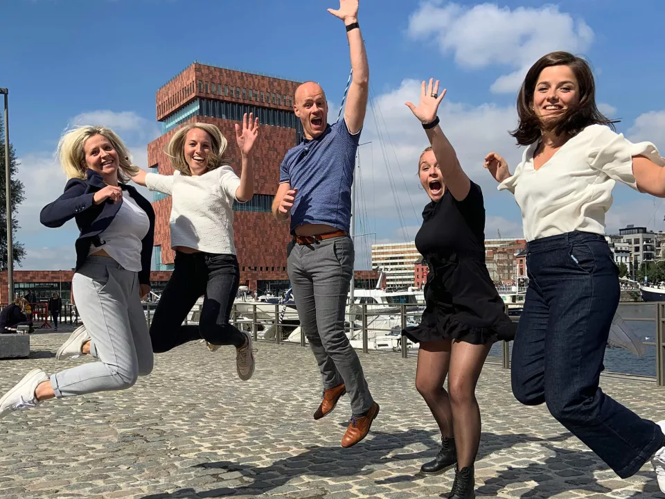 Five people are joyfully jumping on a quay, with a harbour and a modern building in the background.