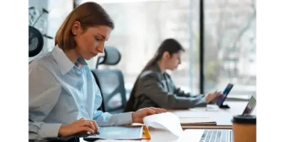 Weibliche Beamtinnen, die in einem Büro mit Computern und Dokumenten arbeiten, verdeutlichen den Bedarf an Transparenz und Modernisierung bei der Zeiterfassung im öffentlichen Dienst.